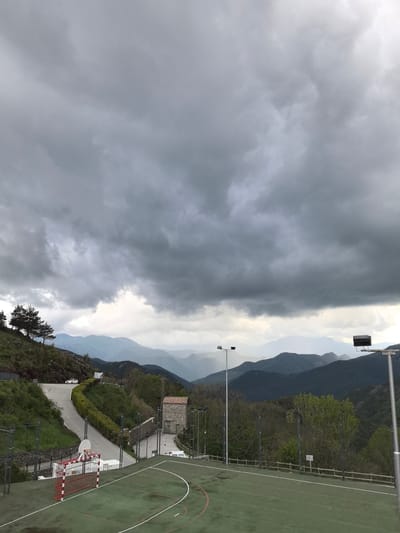 The image depicts an outdoor sports court in Castellar de n'Hug, Spain, under a heavy, overcast sky. The court is green with white lines, and a red and white soccer goal is visible on the left side. A winding road passes behind the court, leading up a hillside with scattered trees and vegetation. In the distance, layers of mountains fade into the horizon. A small stone building sits between the road and the court. Several lampposts are positioned around the court. A white car is visible on the road in the distance.