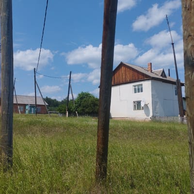 The image depicts a rural residential area on a sunny day with a blue sky and scattered white clouds. The foreground is dominated by tall green grass and two large wooden poles, one on the left and one in the center, obscuring portions of the scene. In the mid-ground, there are two houses and a line of utility poles with wires. The house on the right is a two-story structure with white walls, a wooden-shingled upper gable, a grey roof, and a satellite dish mounted on the lower wall. The house on the left is a single-story building with a brick facade, a grey roof, and a turquoise door. A wooden fence runs along the property line of the house on the right. In the background, there are trees and a distant, partially visible structure. The environment suggests a suburban or village setting, possibly in the city of Kob, Russia, given the context provided.