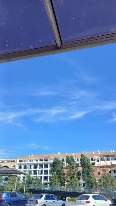The image captures a clear blue sky with wispy white clouds, viewed from under a weathered blue awning with a metal frame. Below the sky and awning, a multi-story building with a terracotta-tiled roof and numerous balconies is visible. Several tall, leafy green trees stand in front of the building. A pedestrian bridge with a metal railing crosses in front of the trees. Several cars are parked or stopped in the foreground, including a blue hatchback, a silver sedan, and a white hatchback. The scene appears to be a street or parking area in a town or city, likely San Pedro De Alcántara, Spain, given the architectural style and sunny weather.