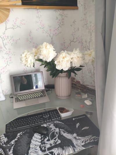 A desk setup is pictured in a room with floral wallpaper. On the desk, there is an open silver laptop with a Cyrillic keyboard, a black keyboard, a black mouse on a dragon-themed mousepad, a white vase with white peonies, and a small white container of food. A light grey curtain is visible on the right side of the image. There is a reflection of a person in the laptop screen. A bamboo fan is positioned in the upper left corner of the image.