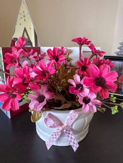 A floral arrangement in a white, heart-shaped vase is centrally located in the image. The flowers are primarily shades of pink and magenta, with dark brown centers. A pink and white checkered ribbon adorns the vase. The arrangement sits on a dark brown surface. In the background, blurred objects and a beige wall are visible. A decorative, triangular object with etched designs can be seen to the left. The image appears to be taken indoors. No information is available regarding the image's location within Rogozinovo, Bulgaria, or the presence of any people or specific actions.