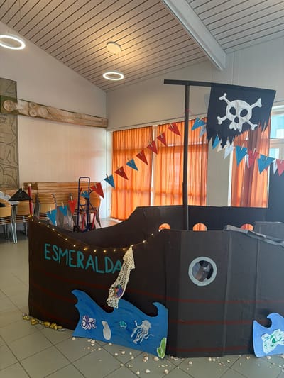 The image depicts a cardboard pirate ship prop indoors, likely in a community hall or school in Ufhusen, Switzerland. The ship is dark brown with red stripes and decorated with a pirate flag featuring a skull and crossbones at the top. A string of fairy lights is draped along the top edge of the ship. The name "ESMERALDA" is written in teal letters on the side of the ship. Below the ship, there are blue cutouts representing waves with illustrated sea creatures such as an octopus and a jellyfish. The floor is tiled in a neutral color and scattered with seashells. In the background, orange curtains are visible, along with stacks of chairs and a blue wheeled cart. Two modern circular pendant lights hang from a sloped, wood-paneled ceiling. A wooden carving of a figure is mounted on the wall to the left. There are no people visible in the image.