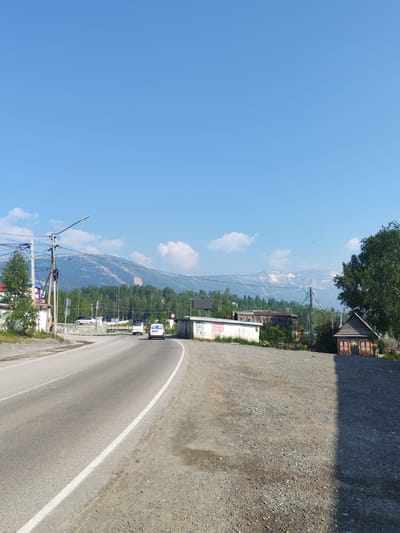 The image depicts a roadside scene in the city of Sheregesh, Russia, on a clear, sunny day. A paved road curves from the left foreground towards the right, bordered by a gravel shoulder. In the distance, a range of mountains dominates the horizon, with some snow visible on their peaks. A dense forest stretches across the lower slopes of the mountains.
Along the roadside, various structures are present. On the left, a white building with some graffiti is visible, adjacent to a large tree. Further down the road, several vehicles are in motion, including a white car and two vans. Electrical poles with wires are scattered throughout the scene.
On the right side of the road, a small wooden structure with colorful murals painted on its facade is prominent. Another dilapidated wooden building is situated behind it. The overall impression is that of a rural or semi-urban environment on the outskirts of Sheregesh, with natural landscapes meeting man-made structures.