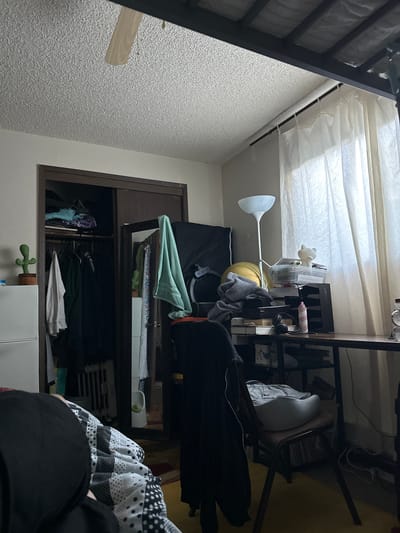 The image depicts a cluttered bedroom. A closet is visible on the left, filled with clothes. A desk area is to the right, with a lamp, various items, and a chair with a pillow on it. A window with sheer curtains is also present. There are no identifiable people in the scene. The location is not discernible from the image.
