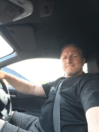 In Quarto, Italy, a middle-aged Caucasian male with light-colored hair and a short beard is seated in the driver's seat of a car, holding the steering wheel. He is wearing a black t-shirt and grey shorts. A grey seatbelt is visible across his chest. The interior of the car, including the dashboard, sun visor, and door panel, is dark. The exterior is bright, suggesting daytime conditions.