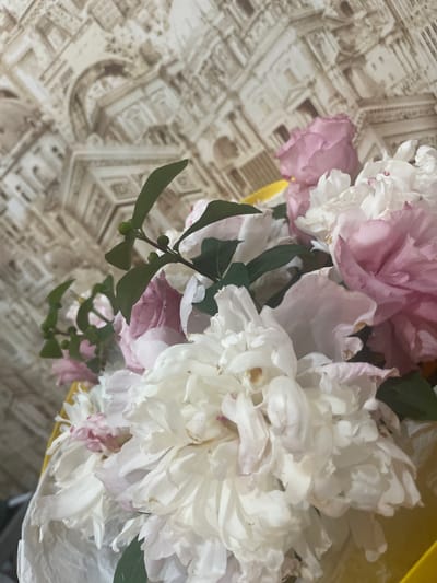 The image displays a close-up of a bouquet of peonies, with a focus on the delicate petals and greenery. The peonies are a mix of white and pale pink hues, with some petals showing slight browning at the edges, indicating they may be past their prime or have been handled. The bouquet is partially wrapped in white paper and held within a yellow container, though the full context of the container is not visible. Delicate green leaves and small, unripe berries are interspersed among the flowers, adding texture and contrast.
The background is a patterned surface with a sepia-toned design that resembles architectural elements or ornate masonry. The pattern is detailed, featuring what appear to be columns, arches, and building facades. This background is out of focus, which helps to draw attention to the flowers.
The lighting appears to be soft and natural, suggesting it might be daytime, though there are no specific time-of-day cues like sunlight direction or shadows. The overall mood is gentle and serene, amplified by the soft colors of the flowers and the blurred, unobtrusive background.
There is no visible text in the image. The scene is likely indoors, given the controlled lighting and the backdrop, though its specific location cannot be determined beyond the provided context of Gukovo, Russia. No people are visible, and the focus is solely on the floral arrangement and its immediate surroundings.
