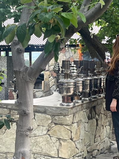 The image shows a display of shiny, metal samovars, possibly for sale or exhibition. A person is taking a photo of the display with their smartphone. The background consists of stonework, suggesting an outdoor setting. A tree with green leaves and branches frames the left side of the image. The image is shot from a ground-level perspective, with a slight upward angle. The location is identified as the city of Miatli, Russia.