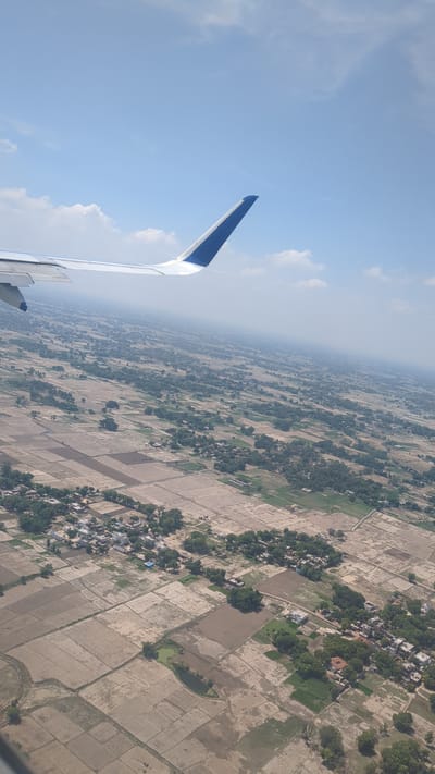 An aerial view captures the landscape of Babatpur, India, from an airplane window. The image is dominated by the airplane wing, which occupies the upper left portion of the frame and features a distinctive blue stripe on its tip. Below, the rural scenery unfolds, characterized by a patchwork of agricultural fields, some appearing fallow with dry soil and others showing signs of cultivation with greener patches. Small clusters of houses, typical of Indian villages, are scattered throughout the landscape, nestled amongst trees. A hazy atmosphere suggests the image was taken during daytime with the sun high in the sky. The overall impression is one of a vast, cultivated rural expanse under a bright, partly cloudy sky.