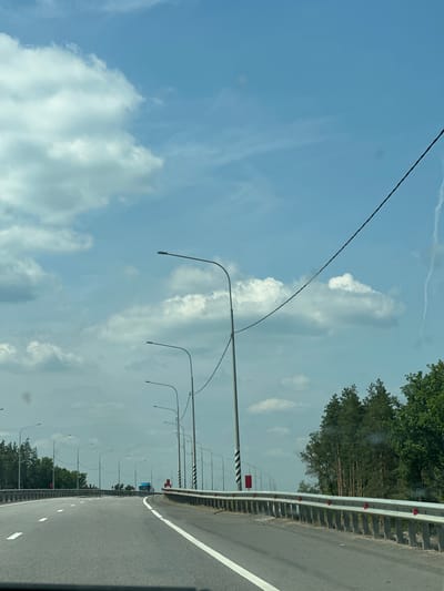 The image captures a forward view from a moving vehicle on a highway, likely a bridge or elevated roadway, given the guardrails and the perspective. The sky is blue with scattered white clouds. A series of streetlights are visible along the right side of the road, extending into the distance. A power line runs diagonally across the upper portion of the frame. On the right, a dense line of coniferous trees forms the edge of the landscape. The highway has clear white lane markings. A blue truck is visible in the distance on the highway. The location is identified as Rasskazovo, Russia.