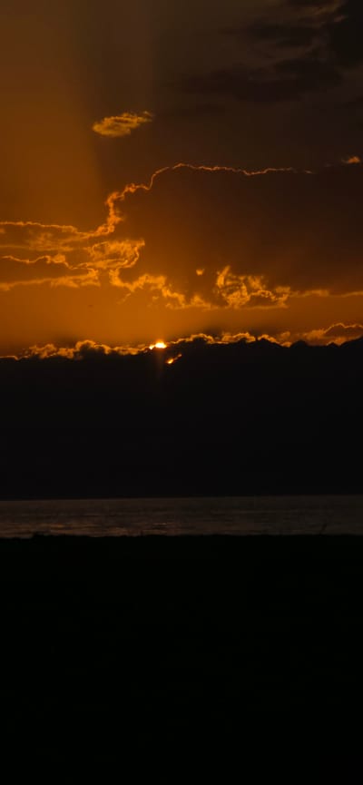 A vertical, high-angle shot captures a dusky sunset over a body of water. The sky is ablaze with the orange and gold hues of sunset, with streaks of light emanating from behind dark, silhouetted clouds. These clouds form a jagged, uneven line across the middle of the frame, partially obscuring the sun. Below the clouds, a dark, hazy band suggests a distant shoreline or landmass. The water in the foreground is a muted gray, reflecting the subdued light of the sky with a subtle shimmer. The bottom of the frame is filled with a deep black silhouette, likely representing foreground land or vegetation, which is featureless and dark. The overall impression is one of dramatic natural beauty, with the vibrant sky contrasting sharply with the dark foreground and water. The image is void of any human presence or man-made structures.