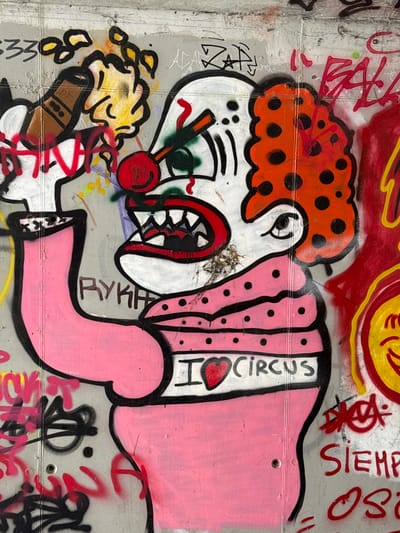 The image is a close-up of a graffiti artwork on a concrete wall in Arenys De Mar, Spain. The artwork depicts a stylized clown or character with exaggerated features. The character has a round, orange head with black polka dots and a white face with a large, open mouth showing sharp teeth and a red tongue. It wears a pink polka-dotted outfit and has a red nose and red and green accents around its eyes. A red heart symbol is visible on the chest, within a white area that also contains black lettering. To the left, another cartoonish figure, possibly a smaller clown or a child, is depicted with yellow hair and holding a brown object, possibly a cigar or a toy. The background is a concrete wall with visible horizontal lines, suggesting it is a building exterior. There are other graffiti tags and splashes of paint in red, yellow, and black scattered around the main artwork.
Visible text in the graffiti includes "I ♡ CIRCUS" written vertically on the character's torso. Above and to the right of the main figure, red spray-painted text reads "SIEMPRE" (Spanish for "always") and possibly other fragmented words. On the lower left, there are black graffiti tags that are difficult to decipher fully but include numbers and abstract shapes. On the lower right, there are more red and black graffiti tags.
The overall impression is vibrant and somewhat chaotic, typical of street art. The time of day and weather are not discernible from the image. The location context is provided as Arenys De Mar, Spain.