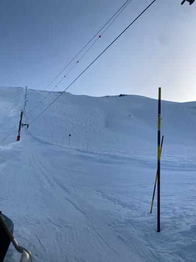 The image displays a snow-covered ski slope in La Salle-les-Alpes, France, under a clear, pale blue sky. The terrain is primarily white snow, exhibiting varied textures.
On the left side of the mid-ground, a section of the ski slope shows distinct parallel corduroy grooming patterns, indicative of recent snowcat activity. To the right, the snow surface appears less uniformly groomed, presenting undulating patterns and scattered tracks.
A ski lift system extends diagonally from the mid-ground up the left side of the slope. This system comprises multiple dark-colored pylons supporting overhead cables. Attached to these cables, at regular intervals, are small, red spherical marker balls.
In the lower right foreground, a thick, vertically oriented black pole with alternating yellow and black stripes is embedded in the snow. A second, thinner pole with a similar yellow and black stripe pattern leans against or is positioned immediately behind the primary pole. Further up the slope, several slender dark poles, some with indistinct markers, are sparsely distributed. A circular dark sign mounted on a pole is visible in the mid-ground, slightly left of center.
The background features steep, snow-clad mountain slopes, with dark rocky outcrops visible along the higher ridges. The scene is illuminated by bright daylight, with a minor atmospheric haze or glare present in the upper right portion of the sky. No individuals are present in the frame. A metallic object, consistent with a shovel blade and part of a wooden handle, is partially visible in the extreme lower-left corner.