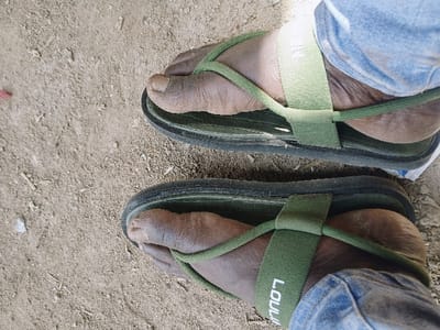 A top-down view displays two human feet, clad in olive green fabric sandals with dark soles, resting on a dry, light brown earthen surface. The individual's skin tone is dark, and the great toenails show minor discoloration. The left sandal's main strap features "LOULUU" printed in white. Light blue denim fabric, consistent with trousers, is visible around the ankles. The ground consists of loose soil with scattered small debris. The scene is located in Mumbwa, Zambia.