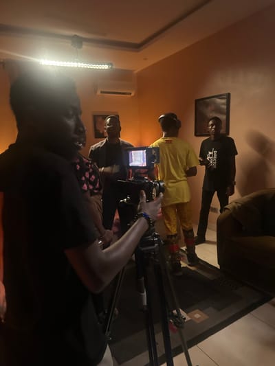 The image shows a film set in an indoor location in Pakuro, Nigeria. A group of individuals are present, seemingly involved in a film production. In the foreground, a person's arm and shoulder are visible, holding a camera mounted on a tripod. The camera is pointed towards the center of the scene.
Further back, several people are visible. One individual, wearing a yellow shirt and patterned pants, stands with headphones on. Another person, dressed in a black t-shirt with "DIRECTOR" printed on it, is also standing. A third person in the background appears to be wearing a suit jacket. A woman in a floral-patterned top is partially visible between the person in the suit and the individual holding the camera.
The environment appears to be a room with a warm-toned paint on the walls. A long fluorescent light fixture is mounted on the ceiling, and an air conditioning unit is visible above a framed picture on the wall. A couch is present in the background to the right. The floor is covered with tiles and a patterned rug. The overall impression is a small-scale film shoot taking place in what seems to be an indoor studio or a well-lit room.