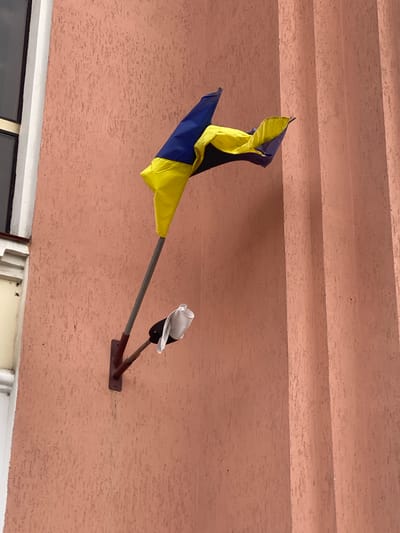 A Ukrainian flag, consisting of blue and yellow horizontal stripes, is affixed to a pole extending from a textured, peach-colored wall. The flag is partially furled and appears somewhat tattered, with its lower section draped over the pole. Attached to the pole below the flag is a smaller, white cloth object, possibly a handkerchief or a piece of paper, also appearing somewhat crumpled. The wall itself has a rough, stucco-like finish and is vertically ribbed by what appear to be architectural columns or decorative elements. To the left of the flag assembly, a portion of a window with white trim is visible, suggesting the flag is mounted on the exterior of a building. The lighting suggests it is daytime, with diffused light casting soft shadows. This scene is located in the city of Stadnia, Ukraine.