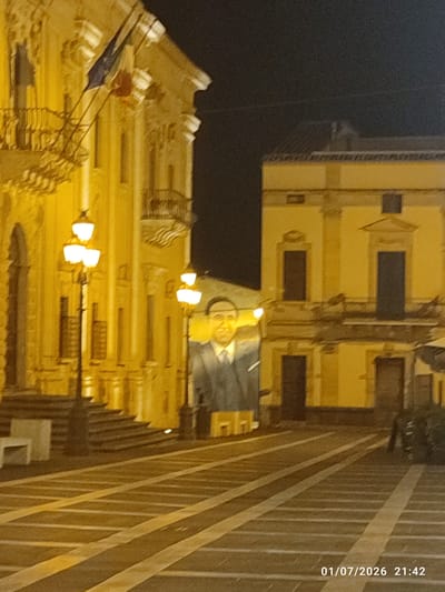 This is a nighttime, medium-range, eye-level shot of a street scene in Militello in Val di Catania, Italy. The image is dominated by the warm glow of streetlights illuminating the facades of two buildings. On the left, a large, ornate building with a balcony and an archway is visible. Two flags, one blue, white, and red and another with red and white stripes, are hanging from the building. A lamppost with three globes stands in front of the building. To the right of this building, a large banner with a portrait of a man in a suit and tie is displayed against the dark night sky. Behind this banner, another building with multiple windows and a balcony is illuminated by the streetlights. The street in the foreground is paved with dark stones and features yellow and white painted lines, possibly indicating parking spaces or a pedestrian crossing. The overall impression is a quiet, festive, or commemorative evening in a historic Italian town.