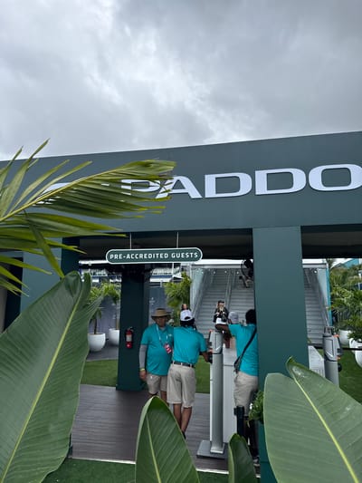 The image shows an outdoor scene at what appears to be an event entrance in Miami Gardens, Florida. A large sign with the word "PADDOCK" in white letters is prominently displayed on a dark gray or green background. A sign below reads "PRE-ACCREDITED GUESTS". Two individuals wearing light-colored shorts and blue shirts are positioned behind a desk-like structure; they may be checking credentials. One of them wears a wide-brimmed hat. Various potted plants, including palms and banana-like trees, are scattered around the area, contributing to a tropical atmosphere. The sky is overcast. The location is suggested by the presence of a Paddock Club, common at auto racing circuits such as the Miami International Autodrome.