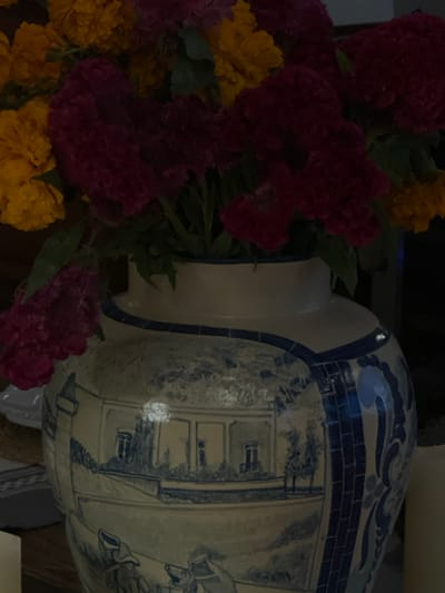 This image, captured in Guanajuato City, Mexico, presents a close-up, eye-level view of a decorative ceramic vase holding a dense floral arrangement. The bouquet consists primarily of ruffled, deep magenta/burgundy cockscomb flowers and bright yellow marigolds, interspersed with green foliage. The vase is a sizable, glazed ceramic object, predominantly white or cream, adorned with intricate blue hand-painted designs. These designs include a continuous horizontal landscape scene depicting multiple colonial-style buildings with windows and balconies, along with stylized trees and small, indistinct human figures in the lower portion. Vertical decorative panels featuring blue geometric or scroll-like patterns are visible on the vase's right side, and its neck and upper shoulder are banded in blue. The scene is dimly lit, with reflections from an unseen light source apparent on the vase's glossy surface. In the lower left foreground, a section of a white, fluted plate and a woven mat are visible. To the lower right, a cylindrical, light-colored object, consistent with a candle, is partially depicted. The background is dark and indistinct. No specific landmark or outdoor scene within Guanajuato City is discernible from the image content.
