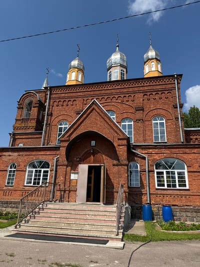 This image depicts the front facade of a red brick church, likely located in Chudovo, Russia, under a clear blue sky with scattered clouds. The church features a bell tower on the left, a central entrance with a large arched doorway and wooden doors, and three onion-shaped domes topped with crosses on the roof. The architecture is characteristic of Eastern Orthodox churches, with ornate brickwork and arched windows.
The entrance is accessed by a wide set of stairs with red risers and stone treads, flanked by metal railings. There is a dark mat at the bottom of the stairs. To the right of the entrance, two large blue plastic barrels are visible, situated on a paved area near a stone wall. Green grass and some flowering plants are growing in the foreground and to the sides of the church. A black hose lies on the ground in front of the barrels. The overall scene suggests a sunny day in a rural or semi-urban setting. There are no people visible in the image. The perspective is from ground level, looking slightly upwards at the church.