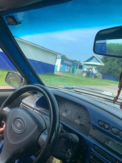 The image is a point-of-view shot from inside a car, looking through the windshield. The car's interior is visible, including the steering wheel with a "PILOT ULTRA" emblem, the dashboard with a speedometer and tachometer, and a blue trim. Outside the car, a rural scene unfolds. Two buildings with blue and white facades are visible, along with a blue tractor. The environment appears to be a village, possibly in Russia, given the architectural style and the presence of a tractor. The sky is clear and blue, suggesting daytime. There are no people clearly visible within the frame, except for a hand holding the steering wheel.