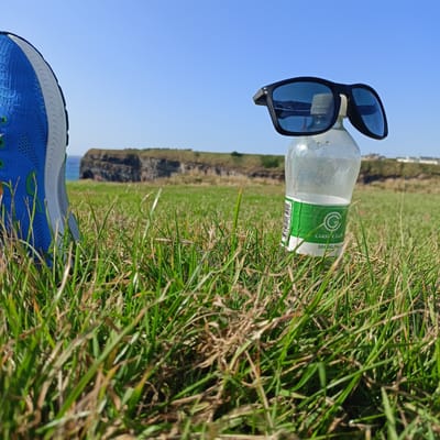 An eye-level medium shot captures a pair of black sunglasses resting on the cap of a clear plastic bottle of sparkling water. The bottle has a green label with the Carrick Glen logo and text that reads "SPARKLING WATER." The scene is set on a grassy field with a low cliff in the background, overlooking the ocean. A blue running shoe is visible in the foreground, to the left of the bottle. The sky is clear and blue. The location is Ballybunnion, Ireland.