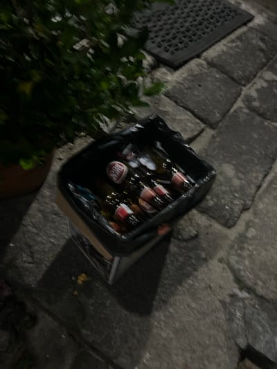 The image shows a black trash bag filled with several glass bottles, primarily beer bottles, placed on a cobblestone pathway. The bottles have red and white labels, with some appearing to be from the brand "Super Bock". A green plant is visible to the left of the trash bag, in a brown pot. A black metal grate is also present in the upper right portion of the image. The scene is outdoors, likely on a street or patio in Felgueiras, Portugal, given the presence of cobblestones and the characteristic plant life and a metal grate often found in Portuguese urban settings. The lighting suggests it is either nighttime or dusk. There are no people or interactions visible in the frame.