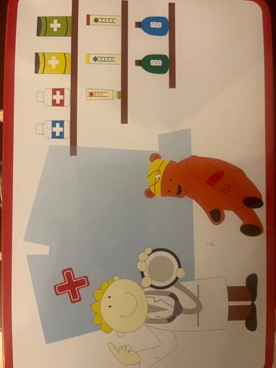 The image shows a cartoon illustration of medical items and characters on a light-colored surface with a red border. On the upper left, there are four rows of medical supplies displayed on brown shelves. The first row has a green rectangular object with a white cross. The second row has a yellow rectangular object with a white cross. The third row has a white bottle with a red cross. The fourth row has a white bottle with a blue cross. To the right of these, there are syringes and vials. The first row has a yellow syringe with blue markings. The second row has a light blue syringe with blue markings. The third row has a red and white striped tube with a green cap. The fourth row has a green vial with a white wavy line.
In the lower portion of the image, a cartoon of an orange bear lies on its side. The bear has a yellow bandage on its head and a red bandage on its arm. To the right of the bear, a cartoon of a doctor is shown. The doctor has yellow hair and is wearing a white coat. The doctor is holding a stethoscope. A large blue shape is behind the doctor and the bear, and a red cross symbol is visible on this blue shape. The image appears to be a playful, child-oriented illustration related to healthcare.
There is no information in the image that would allow for the determination of its location in the city of Edem, Russia, or any other specific geographical location. The scene is an artistic representation, not a real-world setting.