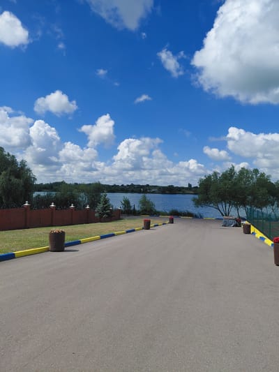 This is a medium shot, taken from a low angle, of a paved pathway leading towards a body of water on a bright, sunny day. The sky is a vibrant blue, filled with fluffy white cumulus clouds. In the foreground, the asphalt pathway is clear, with yellow and blue painted curbs along its edges. Several large, cylindrical planters containing flowers are placed along the left side of the pathway, as well as on the right side closer to the water.

A low, red brick wall with decorative white caps runs along the left side of the pathway, separating it from a grassy area with scattered small white flowers. Beyond the wall, there are trees and dense green bushes.

The pathway opens up to a wide body of water, likely a lake or river, which occupies the middle ground of the image. The water is calm and reflects the blue sky and white clouds. The far bank of the water is lined with trees and foliage, creating a green horizon. On the right side of the pathway, near the water's edge, is a green fence and several trees. A few people can be seen in the distance near the water's edge, and some structures or buildings are visible across the water.

The scene appears to be a public park or recreational area. The location is the park area of the Kovalivka stadium complex in Kovalivka, Ukraine. The image captures a serene and picturesque outdoor environment.