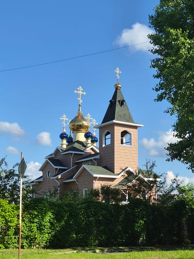 The image shows an Orthodox church in the city of Arseniev, Russia. The church has a light brown exterior with a green roof and multiple domes, one of which is gold and the others are blue. A tall bell tower with a green roof is also visible. The church is surrounded by lush green trees and bushes. In the foreground, there is a patch of green grass and a metal signpost on the left. The sky is blue with some scattered white clouds.