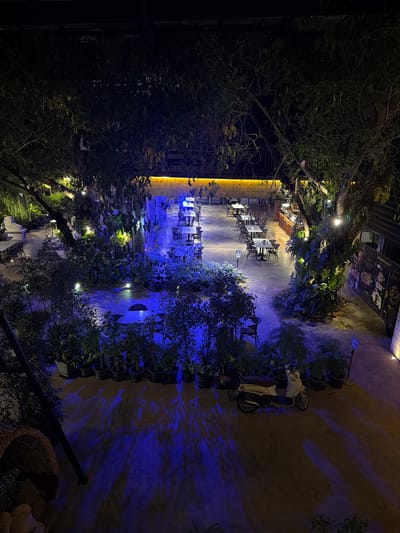 This image depicts an outdoor seating area of a restaurant or cafe at night. The scene is illuminated by artificial lights, including string lights in the trees and a prominent yellow light fixture on a building in the background. The area is furnished with multiple tables and chairs, suggesting it's a place for dining or socializing. Lush greenery, including trees and potted plants, surrounds the seating area, creating an inviting and natural ambiance.
A blue light casts dramatic shadows on the ground and walls, adding a theatrical element to the night scene. In the middle ground, a scooter is parked, and a person is seated on it, partially obscured by shadows and foliage. The overall atmosphere is serene and perhaps a bit mysterious due to the nighttime lighting. The presence of trees and outdoor seating suggests a warm climate.
There are some signs visible on the building in the background, but the text is not clearly legible. The image conveys a sense of a peaceful evening in an outdoor dining establishment. The specific location context provided is Ban Phonsavang Neua, Laos, which aligns with the tropical-like vegetation and outdoor dining setup.