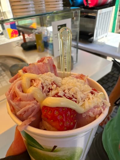 A close-up, eye-level shot shows a person holding a paper cup filled with rolled ice cream. The ice cream is pink and appears to be flavored with strawberries, as evidenced by a whole strawberry on top. It is drizzled with a white, creamy sauce and sprinkled with shredded white pieces, possibly coconut or white chocolate. A translucent yellow plastic utensil is stuck into the ice cream. The cup has a green apple design on it. In the background, a counter is visible with various items, including a red and yellow Red Bull can, a yellow can, and a glass enclosure containing what appears to be a slushy machine. The setting seems to be an outdoor kiosk or stand. The general location is identified as Schönau am Königssee, Germany.