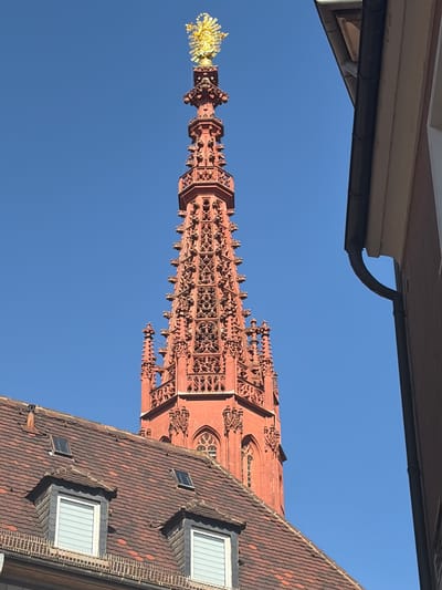 The image shows the tower of the Würzburg Cathedral, also known as the Mariendom, in Würzburg, Germany. The tower is made of red sandstone and is topped with a golden statue of the Virgin Mary. The sky is clear and blue. In the foreground, there is a red tiled roof with dormer windows. A building with a dark downspout is visible to the right of the tower. The image is taken from a low angle, looking up at the tower. The sun is shining, creating strong shadows.