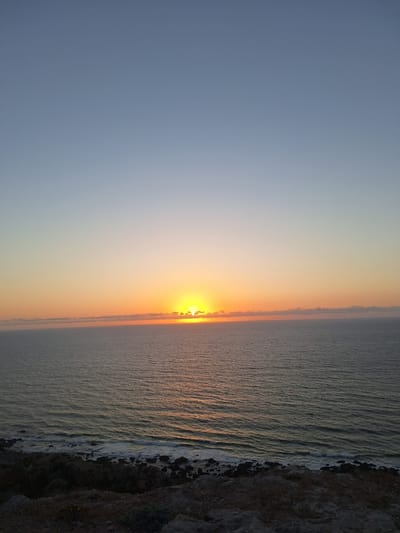 The image shows a sunset over the ocean. The sun is low on the horizon, partially obscured by clouds, casting a warm orange glow across the sky and the water. The ocean is calm with gentle waves breaking on a rocky shore. The foreground is a rocky, vegetated cliff overlooking the sea. The sky transitions from a clear, pale blue at the top to orange and yellow near the horizon. The scene appears to be a natural landscape with no human presence or man-made structures visible. The location is identified as Ahrara, Morocco.