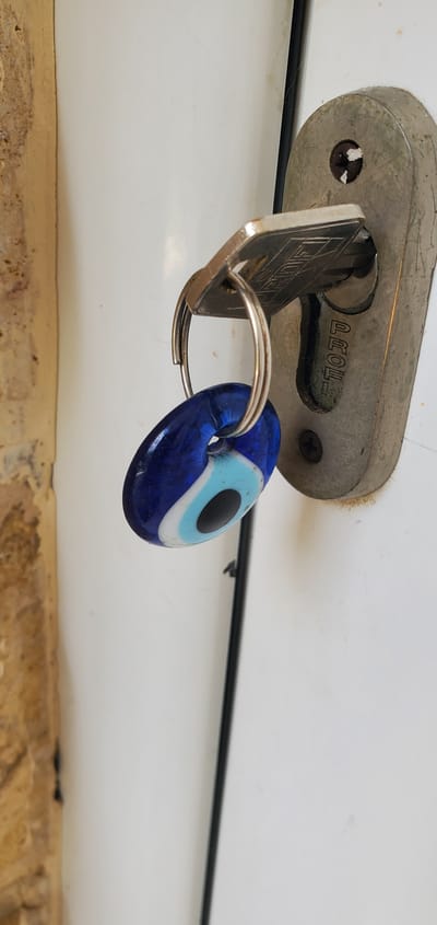In Νέα Σάντα, Greece, a close-up shot focuses on a metallic door lock. A silver key is inserted into the lock, and a keychain with a blue evil eye charm hangs from it. The charm is oval-shaped and displays the characteristic concentric circles of an evil eye, with a dark blue center, surrounded by lighter blue, white, and black rings. The key itself has engraved markings. The lock is mounted on a white door, and to the left of the lock, a textured, beige-colored wall is partially visible. The background is primarily the white door, with a thin black vertical line suggesting a gap or edge. The scene is illuminated, creating subtle shadows and highlighting the textures of the objects.