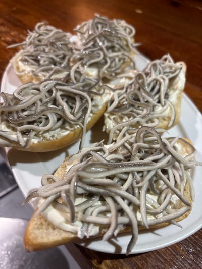 A white ceramic plate, positioned on a dark brown, reflective wooden surface, displays six small bread slices. Each slice is topped with a white creamy spread and a dense cluster of thin, off-white to grayish, noodle-like strands, identified as *gulas* (imitation baby eels) or *angulas* (baby eels). The food items are the sole subjects of the image. No individuals, actions, or specific identifying architectural or environmental features pointing to Piedrahíta, Spain, are discernible within the frame.