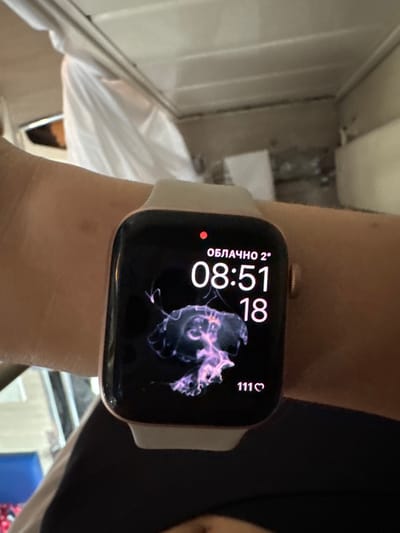The image displays a human left forearm and wrist, with light skin tone, wearing a rose gold-toned Apple Watch with a light-colored silicone band. The watch face is active, showing "ОБЛАЧНО 2°" (Cloudy 2°C) at the top, the time "08:51," the number "18," and a vibrant, translucent purple and pink jellyfish animation in the center. A "111♡" icon is present at the bottom right of the screen. The background is an out-of-focus indoor environment, featuring light-colored overhead panels or structures, and a white material draped over an unseen object on the left, possibly within a vehicle or room in the city of Gryazi, Russia.