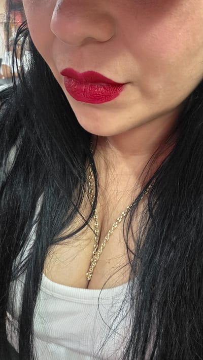 A close-up, cropped, waist-up, outdoor, daytime shot shows a person with fair skin and long, dark hair. The person is wearing a white tank top and a gold chain necklace. Their lips are painted a vibrant red lipstick. The image is a portrait of the person's upper body, with the focus on their lips and décolletage. The background is blurred and indistinct, suggesting an urban environment. The image appears to be taken in Torrejón de Ardoz, Spain.