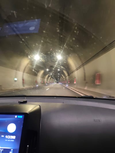 The image shows a car driving through a tunnel. The dashboard is visible, with a digital clock displaying 10:32. The tunnel has lights and emergency exits along the walls. The windshield is slightly dirty. It is the Jeleznitsa tunnel, located in Jeleznitsa, Bulgaria. No people are clearly visible.