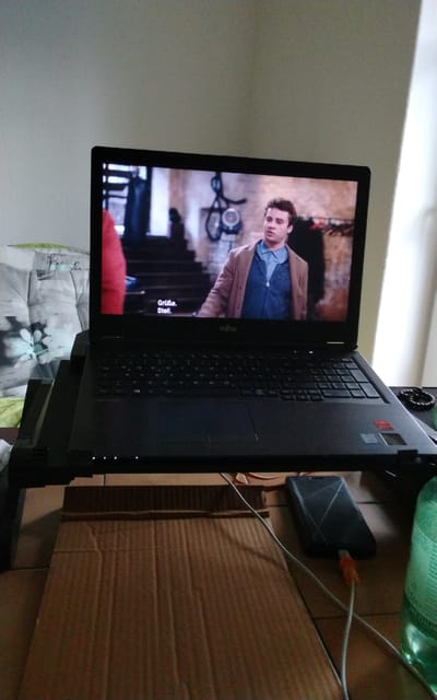 The image shows a laptop placed on a stand, displaying a scene from a movie or TV show. On the screen, a man in a light brown jacket and blue shirt is visible. Subtitles in German, "Grüße. Stell.", are present at the bottom left of the screen. The laptop is on a wooden surface, with a cardboard box underneath it. To the right of the laptop, a smartphone is charging via a USB cable, and a green plastic bottle is also visible. In the background, to the left, a part of a pillow with a floral pattern can be seen. The overall setting appears to be an indoor environment, likely a living room or bedroom. There is no information in the image that suggests a specific location in the city of Halberstadt, Germany.