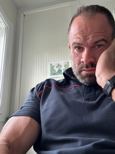 A selfie-style photograph depicts a middle-aged man indoors. He has short, dark hair with a receding hairline, a salt-and-pepper beard, and fair skin. He is wearing a dark-colored polo shirt with red trim, and a black smartwatch on his left wrist. His face exhibits a pensive or slightly tired expression.
The background features a white paneled wall, a window with a white frame, and a printed diagram or map affixed to the wall.
The location is the city of Diekirch, Luxembourg.