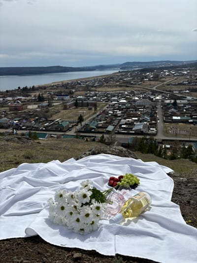 A high-angle, outdoor shot shows a picnic setup on the edge of a cliff overlooking a small town. The picnic consists of a white cloth spread on the ground, on which are arranged a bottle of white wine, two pink-tinted glasses, a cluster of green grapes and strawberries, and a bouquet of white daisies. In the background is the town of Невон, Russia, with a mix of small houses and buildings. Further in the distance are bodies of water and low hills under a partly cloudy sky.