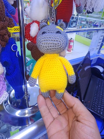 The image shows a close-up of a person's hand holding a crocheted toy hippo. The hippo is grey with a yellow body and grey limbs. It has black button eyes. The toy is attached to a keychain with a silver chain. In the background, there are shelves displaying various other crocheted toys, including a brown bear and a blue octopus. There is also a laptop and a mouse visible on a counter. The scene appears to be in a retail setting, possibly a toy store. The question mentions the city of Ushtobe, Kazakhstan, but there is no visual information in the image to confirm or deny this location. The image is well-lit and the focus is on the toy and the hand.