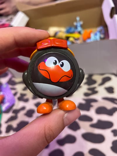 A person's fingers hold a small, black and orange toy penguin with a red helmet. The penguin has a circular body with a cartoonish face featuring large white eyes with black pupils, an orange beak, and a gray chin. The penguin has orange feet and is standing upright. The background is blurry, showing a box with other toys inside, and a surface with a black and white cow-print pattern. There is no information available about the location being Ladozhskii Trudposelok, Russia.