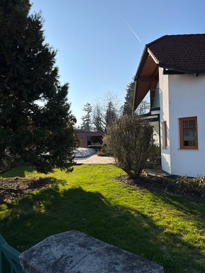 The image depicts a residential scene in Ferschnitz, Austria, under bright daylight with a clear blue sky. A distinct white contrail is visible diagonally across the upper right quadrant of the sky.

In the immediate foreground, a weathered, light-gray stone pillar or cap of a boundary structure is partially visible at the bottom left, with a section of a green metal railing below it. The ground immediately beyond this structure is covered in vibrant green grass, which receives direct sunlight, creating strong shadows from unseen objects to the left.

The midground features a large, dark green coniferous tree on the left, casting a substantial shadow onto the lawn. To the right of the lawn, a deciduous bush with sparse, brown foliage stands near the corner of a building.

In the background, on the right, a multi-story building with white stucco walls and a brown tiled gabled roof is prominent. It features wooden eaves, a small balcony with a glass railing on its upper level, and a rectangular window with a light brown wooden frame on the lower visible section. Sunlight illuminates the right side of this structure. Further left, behind the main coniferous tree, an outdoor patio area is visible, paved with light-colored, irregular flagstones. On this patio, a translucent, dome-shaped cover, consistent with a pool enclosure, is present. Behind the patio, a modern structure with dark paneling and reflective windows is partially obscured by trees. Scattered throughout the background are various trees, some coniferous and others deciduous, largely bare of leaves, suggesting a cooler season. No individuals or active subjects are present.