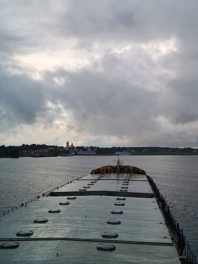 The image depicts a wide river with a barge in the foreground, viewed from its stern. The barge's deck is a metallic, flat surface with numerous circular hatches. A railing runs along the starboard side of the barge. In the distance, on the left bank of the river, lies the city of Myshkin, Russia. The city's skyline features a prominent cluster of buildings with golden domes, likely a church or monastery, and other structures. The water appears calm with gentle ripples. The sky is overcast, with dramatic, dark gray clouds interspersed with patches of brighter, lighter sky. The overall atmosphere is subdued and somewhat moody due to the cloudy weather.