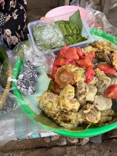 A close-up, overhead shot captures a green plastic tray filled with various Indonesian snacks. The tray, resting on a rough, grey surface, is partially obscured by a clear plastic sheet. To the left of the tray, a person wearing a black dress with a floral print is visible.
The tray's contents include:
* **Green snacks:** A clear plastic container holds several small, green, rectangular snacks, possibly wrapped in banana leaves.
* **Red and golden-brown fried items:** A large portion of the tray is covered with a mix of fried snacks. Some are bright red, while others are golden-brown and appear to be pieces of battered tempeh or other fried items.
* **Wrapped dark items:** To the left of the fried snacks, three small, dark items are individually wrapped in clear plastic, secured with white ties. Their contents are not clearly discernible.
* **Banana leaves:** Some of the snacks are presented on or wrapped in banana leaves.
* **Green cylindrical objects:** Partially visible on the far left are some bright green, cylindrical objects, possibly vegetables or another type of snack.
The overall impression is that of a street food vendor's display in Tambakreja, Indonesia, showcasing a variety of popular local snacks. The lighting suggests an outdoor environment, possibly a market or roadside stall.