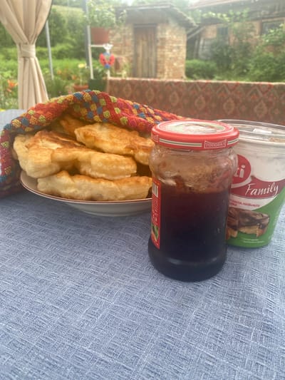This image, captured in Kipilovo, Bulgaria, presents an outdoor scene featuring a collection of food items on a light blue, textured, woven tablecloth. In the foreground, slightly to the left, a white bowl with a thin red-brown rim is filled with numerous pieces of golden-brown, irregularly shaped fried dough, partially covered by a brightly colored, woven textile displaying a checkered pattern of red, orange, green, blue, and yellow squares. To the right of the bowl, a transparent glass jar with a red lid, labeled "TODORKA," contains a dark red substance consistent with jam or preserves. Adjacent to the jam jar, a white and green plastic tub, labeled "Suti Family" and specifying "COCOA, HAZELNUTS & MILK," holds a chocolate spread; a "Serving suggestion" graphic depicting a chocolate-coated pastry is visible on its side.

The background, largely out of focus, reveals an outdoor environment. On the far left, a light beige fabric curtain or drape is tied back. The central background features dense green foliage, suggesting a garden or natural setting, with several potted plants visible. A small, red and black ladybug ornament is perched on a white stand among the greenery. Further back, a patterned textile, possibly a rug or tapestry with red and brown hues, is draped over an unseen object. In the distant background, blurred structures, likely traditional buildings constructed from brick or stone, are discernible through the vegetation, contributing to a rustic atmosphere. No individuals are visible in the scene.