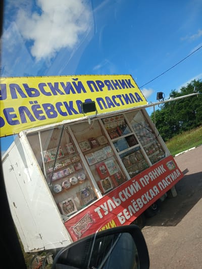 A white, single-axle trailer displays a food stand selling baked goods. The trailer is angled, with its left side towards the viewer. A bright yellow banner with large, dark blue Cyrillic text runs across the top front of the stand. Below this, a glass-enclosed display case reveals shelves stocked with various packaged sweets, primarily circular and hexagonal boxes. Some boxes feature illustrations of people in historical attire. A red banner with white Cyrillic text is stretched across the lower front of the stand. The background consists of a clear blue sky with scattered white clouds, green trees, and a grassy verge alongside an asphalt road. A black car's side mirror is partially visible in the bottom left corner, reflecting the sky. The trailer is parked on a paved surface, possibly a roadside. The specific location is Bol'shie Ploty, Russia.