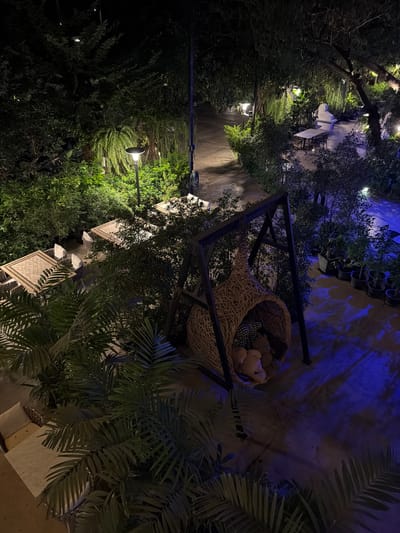 The image depicts an outdoor seating area at night, possibly a cafe or restaurant, in Ban Phonsavang Neua, Laos. The scene is illuminated by artificial lighting, including a street lamp casting a warm glow on lush greenery and a dark pathway. Blue light from a different source casts shadows on a textured wall and plants in pots.
In the foreground, a large woven basket, possibly for decoration, is filled with several light-colored teddy bears. The basket is supported by a dark metal frame. To the right and left of the basket, palm fronds and other dense foliage create a natural, almost jungle-like atmosphere.
Further into the scene, there are glimpses of outdoor furniture. Several small square tables with mosaic tile tops and white chairs are visible, suggesting a place for patrons to relax. The overall impression is one of a tranquil, dimly lit outdoor space, ideal for evening gatherings. The time of day is clearly night, and the weather appears calm, with no indication of rain or wind. There is no visible text in the image.