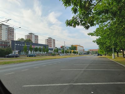 This image captures a street scene in Most, Czechia. The foreground shows a paved road with white lane markings and a parking lot with marked spaces. In the middle ground, a road runs parallel to a grassy median with tram tracks. Several cars are visible on the road and in the parking lot, including a dark sedan and a dark SUV.
Behind the road, a modern building with large windows and several trees line the street. Further in the background, several tall apartment buildings stand against a sky with scattered clouds. The trees in the foreground, with their lush green leaves, suggest it is daytime during a warm season. The sky is a bright blue with wispy white clouds, indicating fair weather. The overall atmosphere of the scene is calm and ordinary, depicting a typical urban environment. No people are clearly visible in the image. There is no visible text in the image.